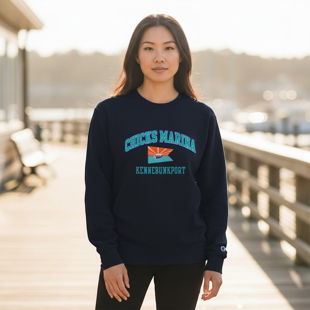 Woman wearing a navy blue Chicks Marina crew sweatshirt with a front logo on a wooden dock at sunset.