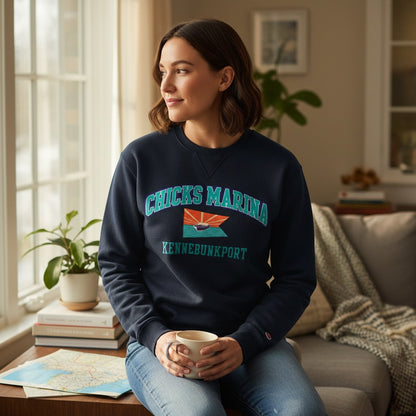 Woman wearing a navy Chicks Marina crew sweatshirt with a graphic logo, sitting by a window in a cozy home setting.